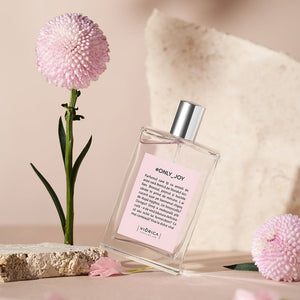 Sticlă de parfum #Only_Joy cu capac metalic argintiu, etichetă roz, flori roz și fundal neutru.