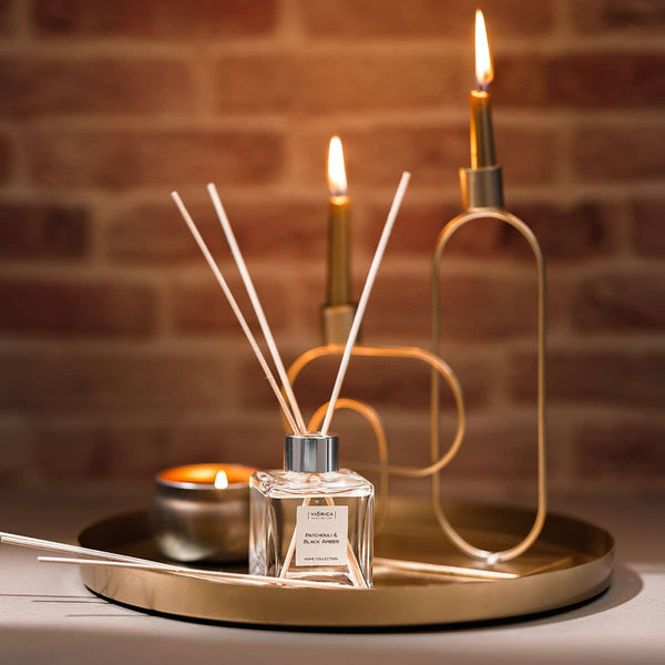 Difuzor parfum patchouli chihlimbar negru pe tavă aurie cu bețe și lumânare în fundal.