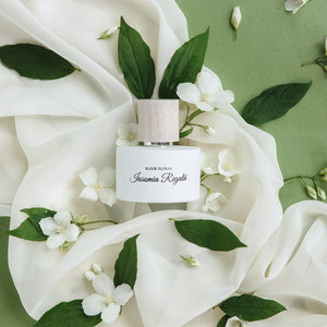 Parfum „Elixir Floral Iasomie Regală”, sticlă albă cu capac din lemn, pe fundal verde cu flori de iasomie.