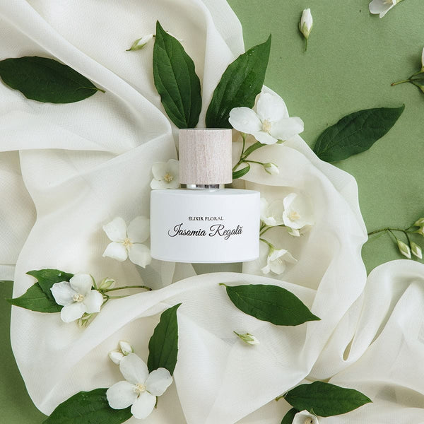 Parfum „Elixir Floral Iasomie Regală”, sticlă albă cu capac din lemn, pe fundal verde cu flori de iasomie.
