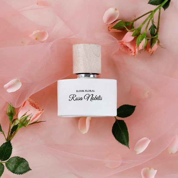Parfum „Elixir Floral Rosa Nobilis”, sticlă albă, capac din lemn, decorat cu petale roz și trandafiri pe fundal de voal roz.