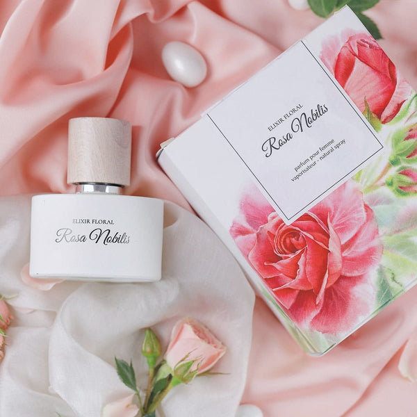Sticluță parfum Elixir Floral Rosa Nobilis cu capac din lemn pe material roz, alături de cutie cu imprimeu floral.