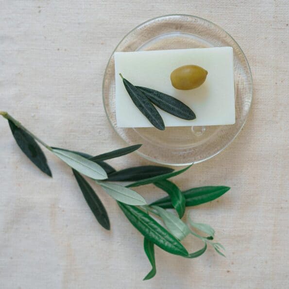 Săpun natural handmade cu măsline pe suport transparent, decorat cu frunze și măslină verde.
